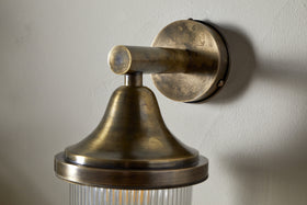 The nkuku Magola Outdoor Wall Lamp in antique brass features a round base, cylindrical arm, flared top, and ribbed glass shade, casting subtle shadows for stylish exterior lighting.