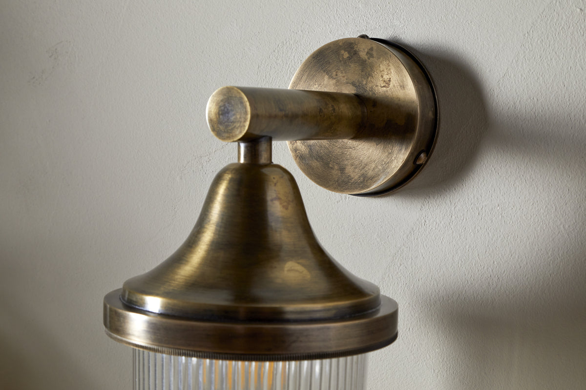 The nkuku Magola Outdoor Wall Lamp in antique brass features a round base, cylindrical arm, flared top, and ribbed glass shade, casting subtle shadows for stylish exterior lighting.
