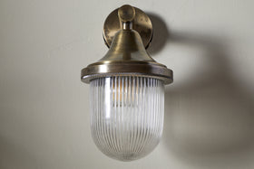 The nkuku Magola Outdoor Wall Lamp in antique brass features a round base, cone top, and ribbed clear glass dome shade. Its vintage-inspired design suits both outdoor and indoor spaces, shown unlit on a light-colored wall.