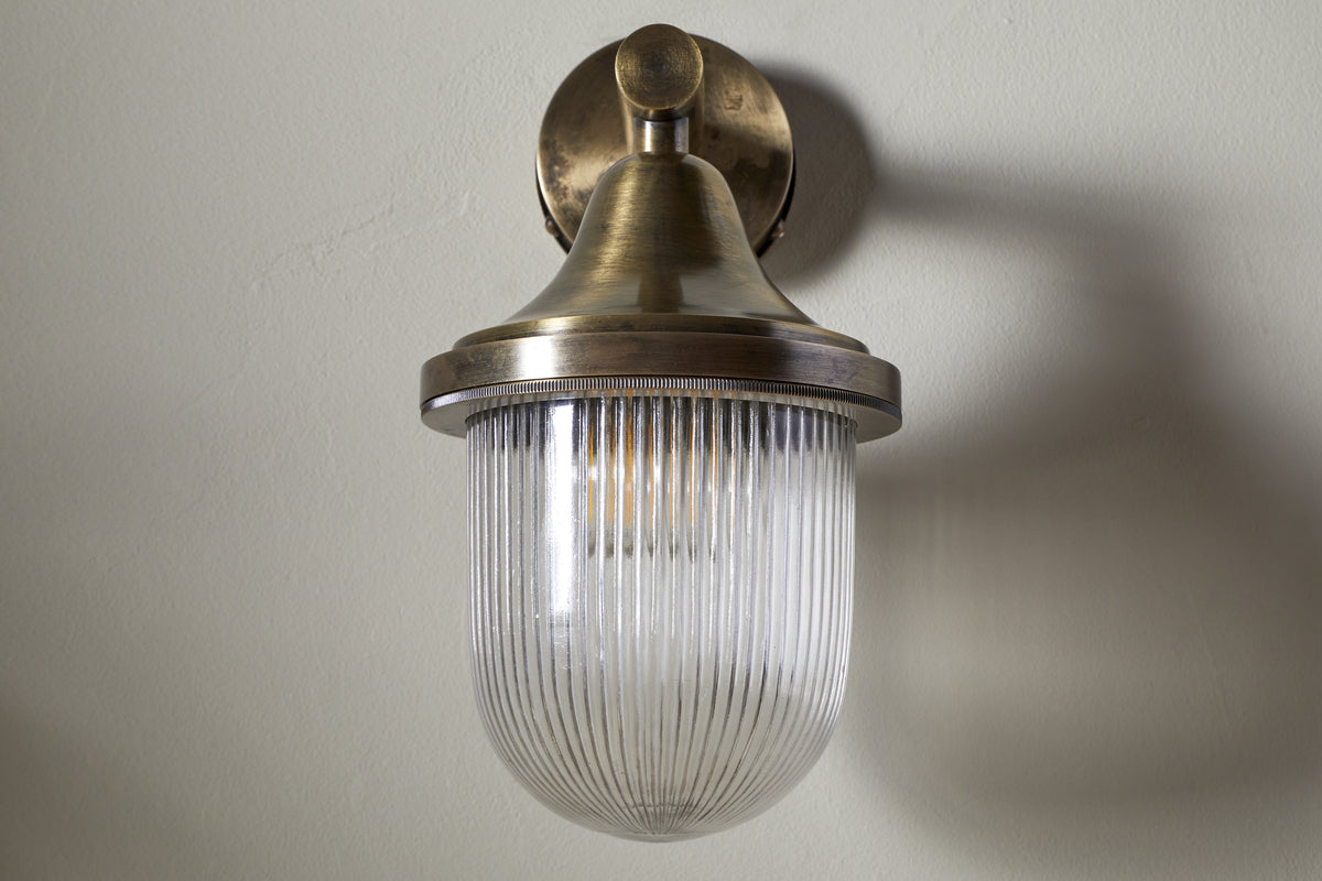 The nkuku Magola Outdoor Wall Lamp in antique brass features a round base, cone top, and ribbed clear glass dome shade. Its vintage-inspired design suits both outdoor and indoor spaces, shown unlit on a light-colored wall.