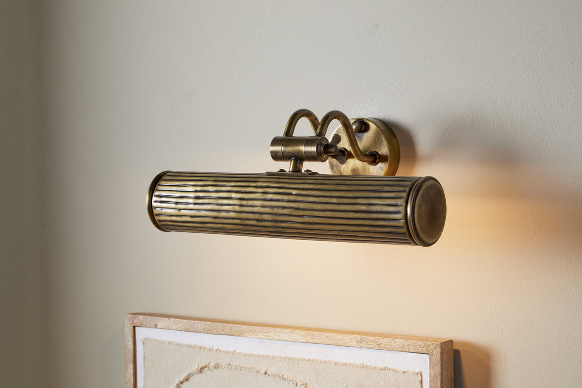 The Tinje Picture Wall Light by nkuku features an antique brass finish and a ridged, cylindrical shade, casting warm downward light onto artwork. Its curved support arms complement neutral walls for elegant display lighting.
