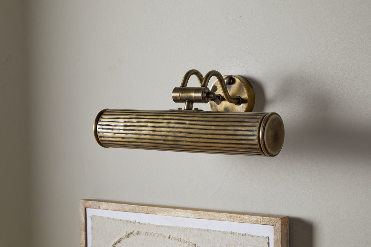 The Tinje Picture Wall Light by nkuku, in antique brass with horizontal ridges and an arched bracket, is mounted above a framed, textured off-white canvas, adding elegant vintage lighting to artwork.