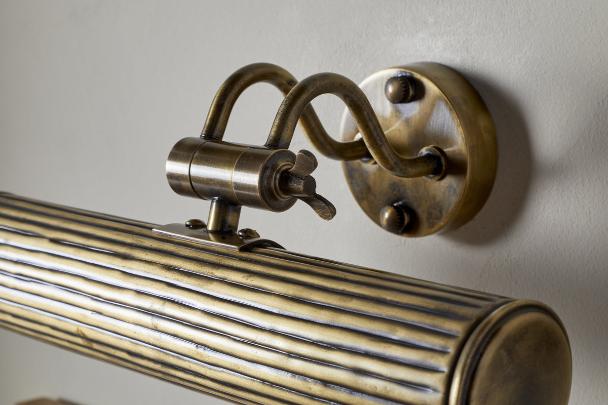 Close-up of nkuku’s Tinje Picture Wall Light in antique brass, featuring a ribbed cylindrical shade, ornate curved arm, and round backplate on a pale wall—perfect for adding elegant lighting to your artwork.