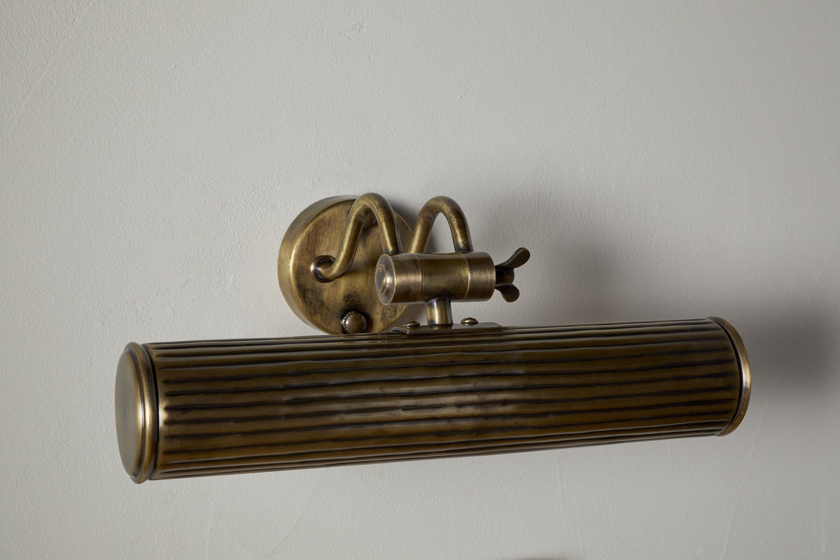 The Tinje Picture Wall Light - Antique Brass by nkuku features a horizontally ribbed cylindrical shade, adjustable arm, and round backplate. Its aged brass finish adds vintage charm, perfect for showcasing artwork on light gray walls.