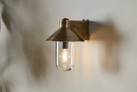 The nkuku Singa Outdoor Wall Lamp in antique brass features a wide, angled shade and clear glass, encasing a glowing bulb that casts warm light and soft shadows on the nearby white wall.