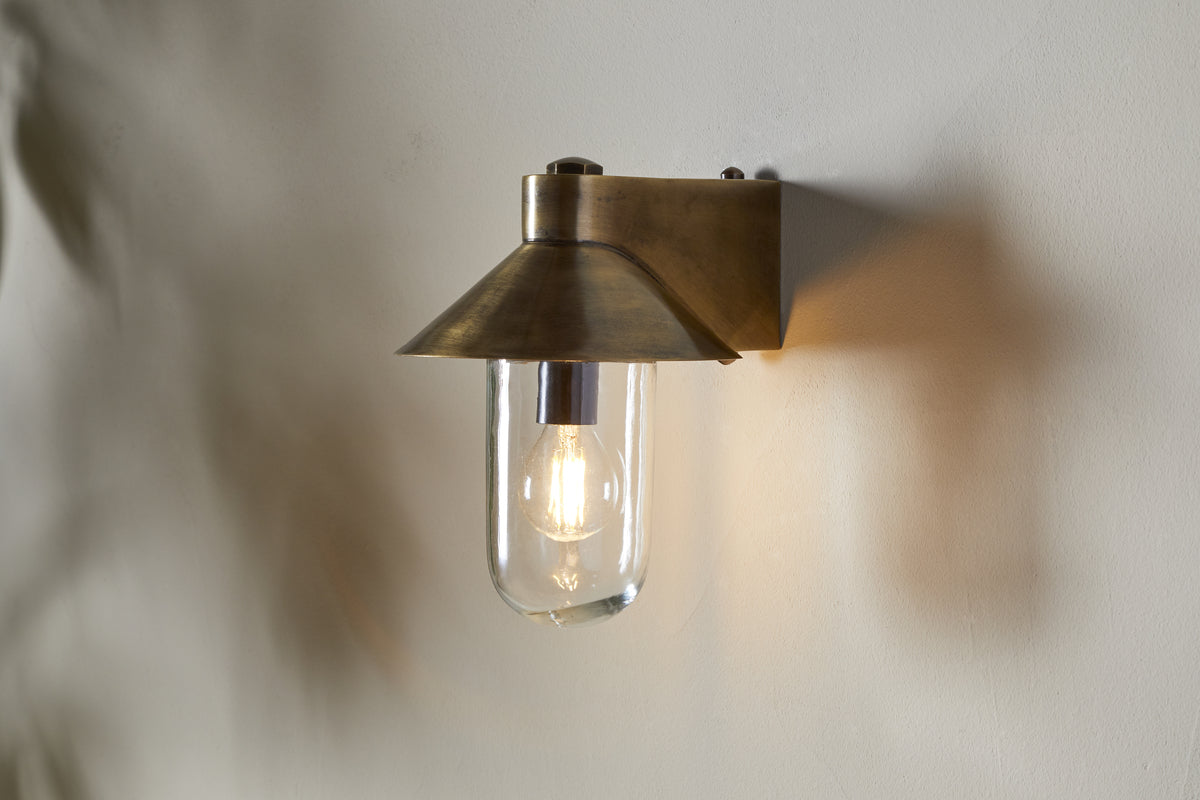 The nkuku Singa Outdoor Wall Lamp in antique brass features a wide, angled shade and clear glass, encasing a glowing bulb that casts warm light and soft shadows on the nearby white wall.