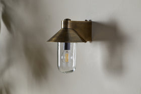 The Singa Outdoor Wall Lamp by nkuku, in antique brass, features a flared conical shade and clear glass that displays an exposed LED filament bulb, casting a soft shadow on the light-colored wall behind it.