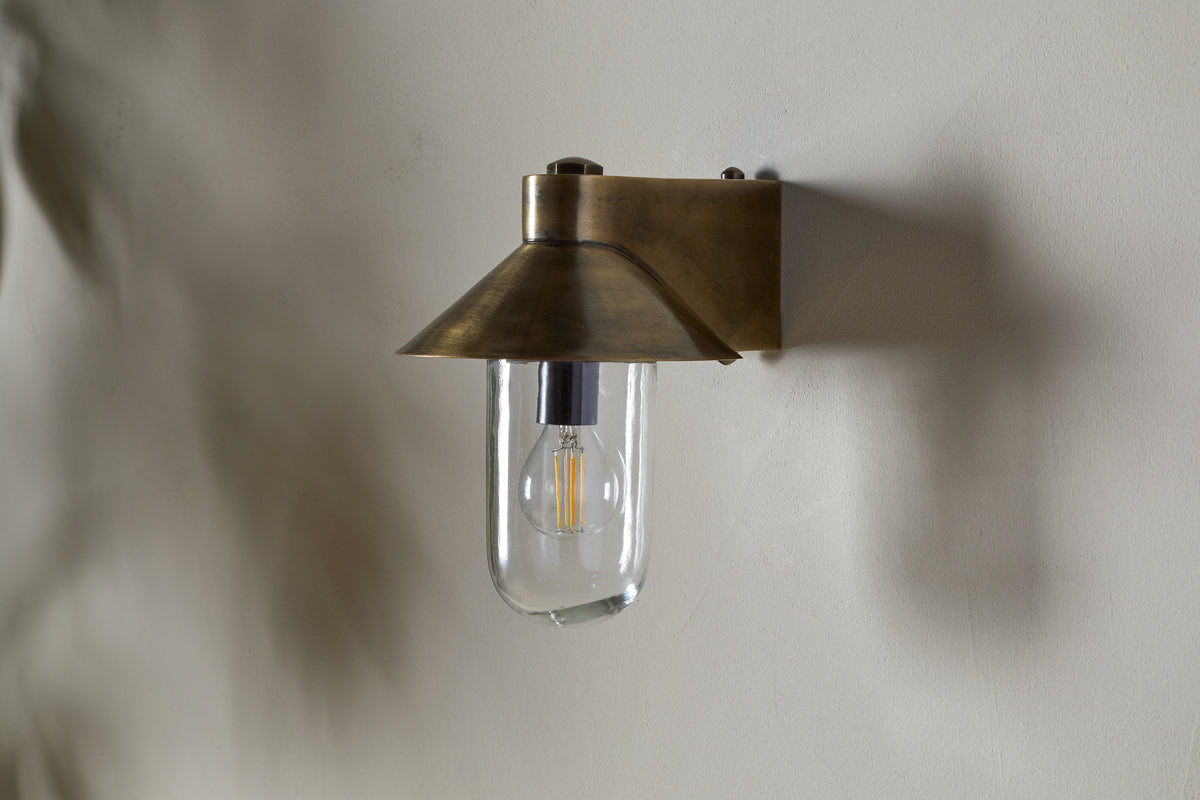 The Singa Outdoor Wall Lamp by nkuku, in antique brass, features a flared conical shade and clear glass that displays an exposed LED filament bulb, casting a soft shadow on the light-colored wall behind it.