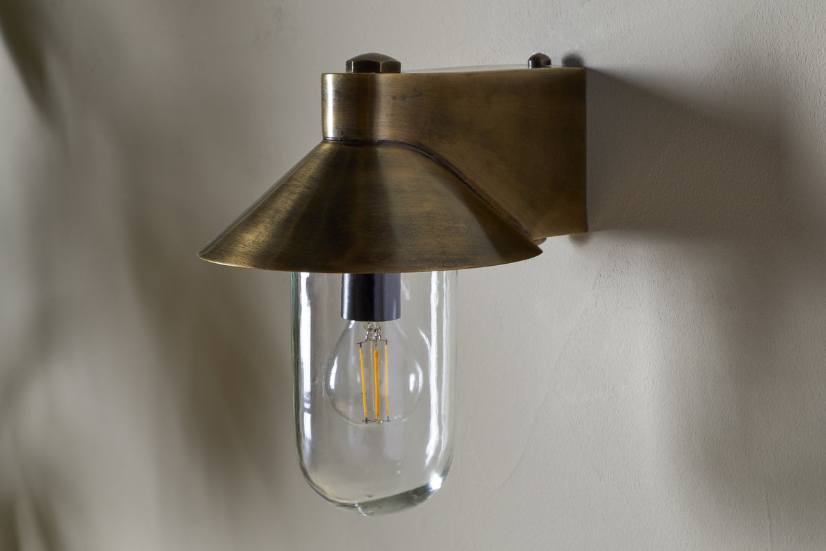 The Singa Outdoor Wall Lamp by nkuku features an exposed LED filament bulb beneath a wide, flared clear glass shade in antique brass. This vintage-industrial wall light boasts a warm metallic finish, perfect for outdoor spaces.