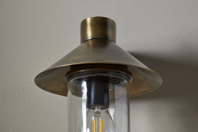 A close-up of the Singa Outdoor Wall Lamp by nkuku in antique brass, featuring a wide conical shade, clear cylindrical glass housing, and visible filament bulb, mounted on a light-colored wall.
