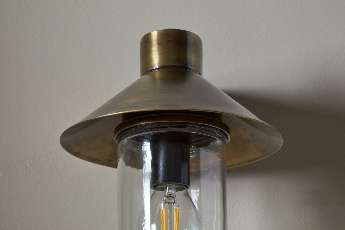 A close-up of the Singa Outdoor Wall Lamp by nkuku in antique brass, featuring a wide conical shade, clear cylindrical glass housing, and visible filament bulb, mounted on a light-colored wall.