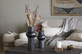A cozy living room with a beige sofa, textured pillows, and a gray throw. On a dark coffee table sits nkuku’s Endo Reclaimed Iron Vase in Aged Black with dried flowers, a white candle on wood, and a cup. A woven basket and tapestry add warmth.