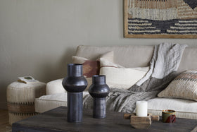 A cozy living room with a beige sofa, gray throw, neutral pillows, woven ottoman with a book, dark coffee table holding an aged bronze vase and the nkuku Endo Reclaimed Iron Vase - Aged Black, two black ceramic vases, a lit white candle, mug, and earth-toned wall tapestry.