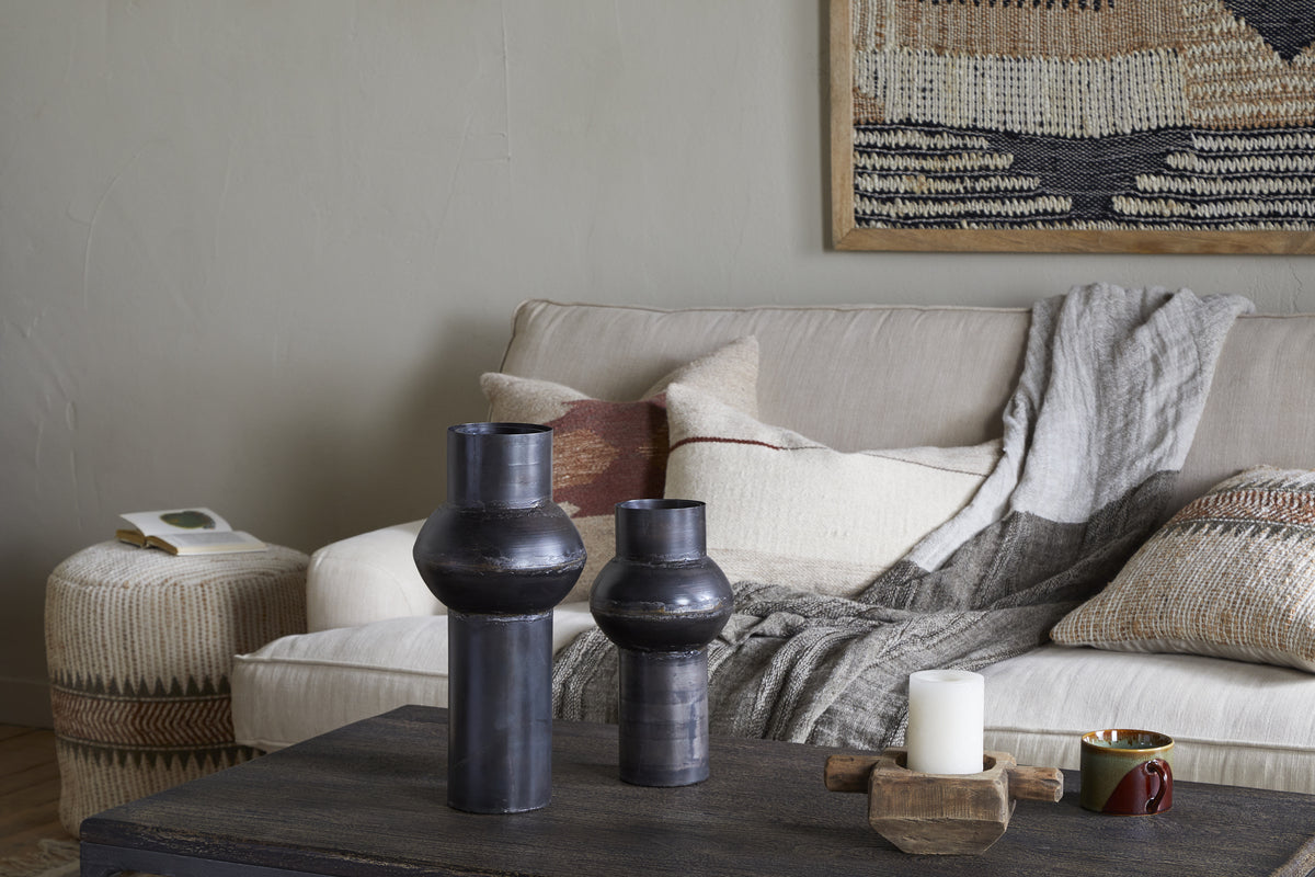 A cozy living room with a beige sofa, gray throw, neutral pillows, woven ottoman with a book, dark coffee table holding an aged bronze vase and the nkuku Endo Reclaimed Iron Vase - Aged Black, two black ceramic vases, a lit white candle, mug, and earth-toned wall tapestry.