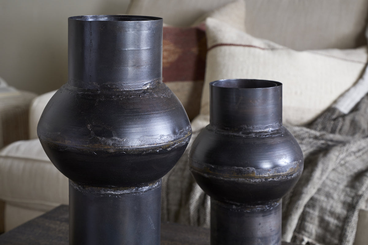 Two nkuku Endo Reclaimed Iron Vases in aged black, featuring a dark matte finish, sculptural shape, and visible welded seams, sit on a table. Behind them is a beige sofa with textured and patterned cushions.