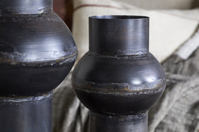 Close-up of two nkuku Endo Reclaimed Iron Vases in aged black, their sculptural forms and visible seams add artisan charm. The rich bronze-black tones complement neutral cushions and a textured gray blanket for a cozy, rustic vibe.