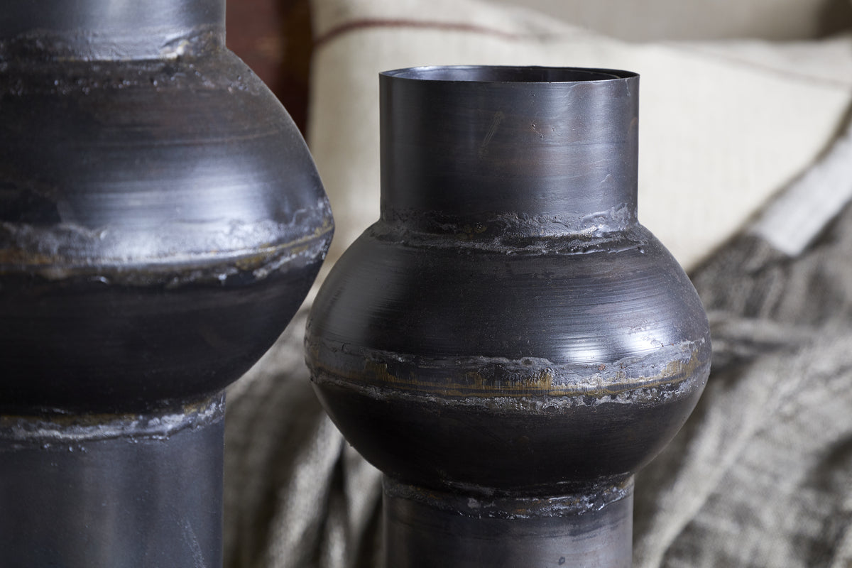 Close-up of two nkuku Endo Reclaimed Iron Vases in aged black, their sculptural forms and visible seams add artisan charm. The rich bronze-black tones complement neutral cushions and a textured gray blanket for a cozy, rustic vibe.