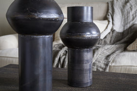 Two Endo Reclaimed Iron Vases in aged black by nkuku, with cylindrical bases and rounded forms, sit on a dark wooden table. A cream sofa with textured pillows and a gray throw blanket creates a cozy living room atmosphere.