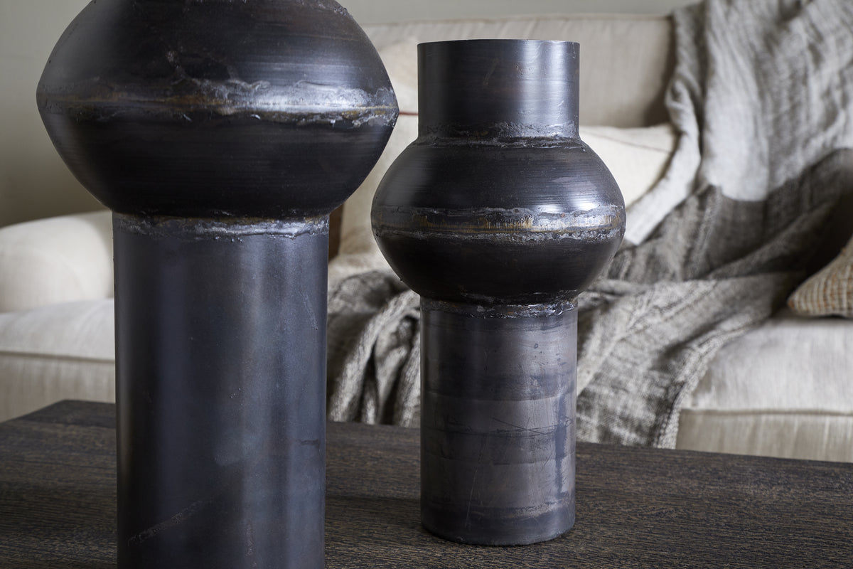 Two Endo Reclaimed Iron Vases in aged black by nkuku, with cylindrical bases and rounded forms, sit on a dark wooden table. A cream sofa with textured pillows and a gray throw blanket creates a cozy living room atmosphere.