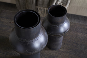 Two Endo Reclaimed Iron Vases in Aged Black by nkuku, with matte finishes and visible welds, stand on a textured brown wooden surface. Their cylindrical forms and wide necks create an industrial, rustic look, with blurred woven fabric behind.