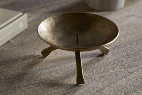 The Kalik Candle Holder by nkuku, crafted in antique brass with three angled legs and a central spike for securing a pillar candle, stands on a textured wooden surface near a beige book and a striped cylindrical object.