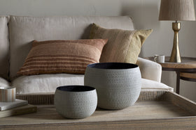 Two nkuku Laoko Olive Grey planters with a textured, round design rest on a wooden tray on the coffee table. Behind them is a cream sofa with striped earthy-toned pillows. A ceramic mug and beige lamp adorn the side table. Perfect for modern spaces.