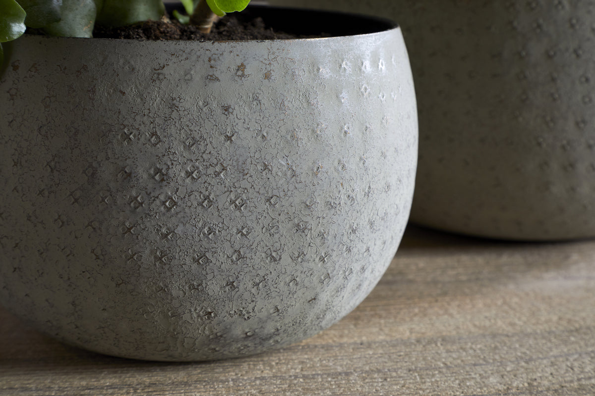 The nkuku Laoko Planter in Olive Grey features a textured, dotted surface and round shape, ideal for modern interiors. A green plant peeks from the top left, with another similar indoor planter softly blurred in the background.
