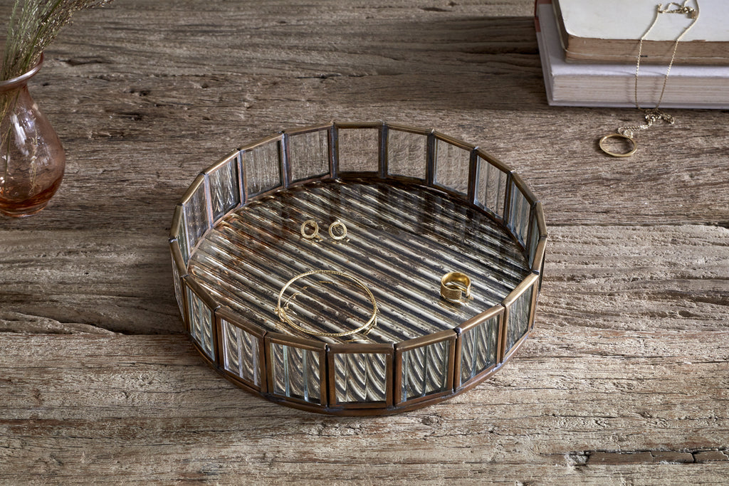 The nkuku Ruaka Glass Vanity Tray in brass sits on a rustic wooden table, displaying gold jewelry—a bracelet, ring, and stud earrings. In the background are stacked books draped with a necklace and a vase of dried flowers.