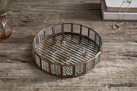 The nkuku Ruaka Glass Vanity Tray in brass sits on a rustic wooden table, displaying gold jewelry—a bracelet, ring, and stud earrings. In the background are stacked books draped with a necklace and a vase of dried flowers.