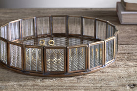 The nkuku Ruaka Glass Vanity Tray – Brass, featuring vertical glass panels in a bronze metal frame, sits on a rustic wooden surface holding gold rings and bracelets, with two stacked books softly lit in the background.