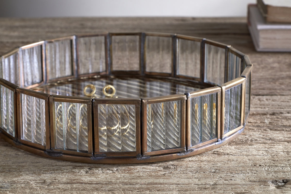 The nkuku Ruaka Glass Vanity Tray – Brass, featuring vertical glass panels in a bronze metal frame, sits on a rustic wooden surface holding gold rings and bracelets, with two stacked books softly lit in the background.
