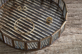 The nkuku Ruaka Glass Vanity Tray - Brass features a round, metallic design with raised edges and a striped, antique mercury glass surface, beautifully displaying gold jewelry on weathered wooden planks.