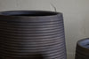 Close-up of the nkuku Lmaga Metal Grooved Planter in Aged Black, featuring textured horizontal lines. The large pot stands before a rough beige wall, with part of a smaller matching planter visible to the right.
