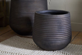 The nkuku Lmaga Metal Grooved Planter in Aged Black stands on a light woven rug atop a wooden floor near a wall, ideal for displaying houseplants. Another similar indoor planter is partially visible in the softly lit background.