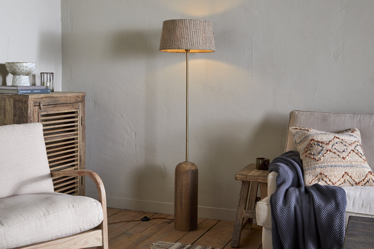 A cozy living room features the nkuku Agua Grooved Floor Lamp - Natural glowing in the corner, with sustainable mango wood furniture, a beige sofa, blue throw, armchair, and textured light walls enhancing the wooden floor.