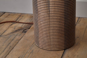 A close-up of the nkuku Agua Grooved Floor Lamp - Natural, crafted from sustainable mango wood, stands on a wooden floor with visible grains. Its brown cord extends behind it—an ideal accent for modern living spaces.