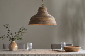 The Nadi Grooved Wood Pendant - Natural & Antique Brass by nkuku hangs above a neutral dining table set with a beige tablecloth, wooden bowl, ceramic vase of green branches, water glasses, plates, and gray upholstered chairs.