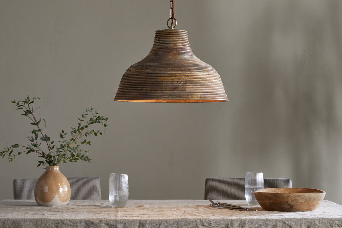 The Nadi Grooved Wood Pendant - Natural & Antique Brass by nkuku hangs above a neutral dining table set with a beige tablecloth, wooden bowl, ceramic vase of green branches, water glasses, plates, and gray upholstered chairs.