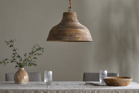 The Nadi Grooved Wood Pendant by nkuku in natural and antique brass hangs above a linen-covered dining table adorned with a beige vase of green branches, ribbed glasses, a wooden bowl, and two forks, set against a light gray wall.