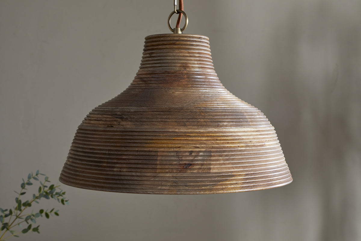 The nkuku Nadi Grooved Wood Pendant in Natural & Antique Brass hangs from a metal chain, its ribbed, bell-shaped mango wood design casting warm light. A small leafy plant appears in the lower left against a blurred neutral background.
