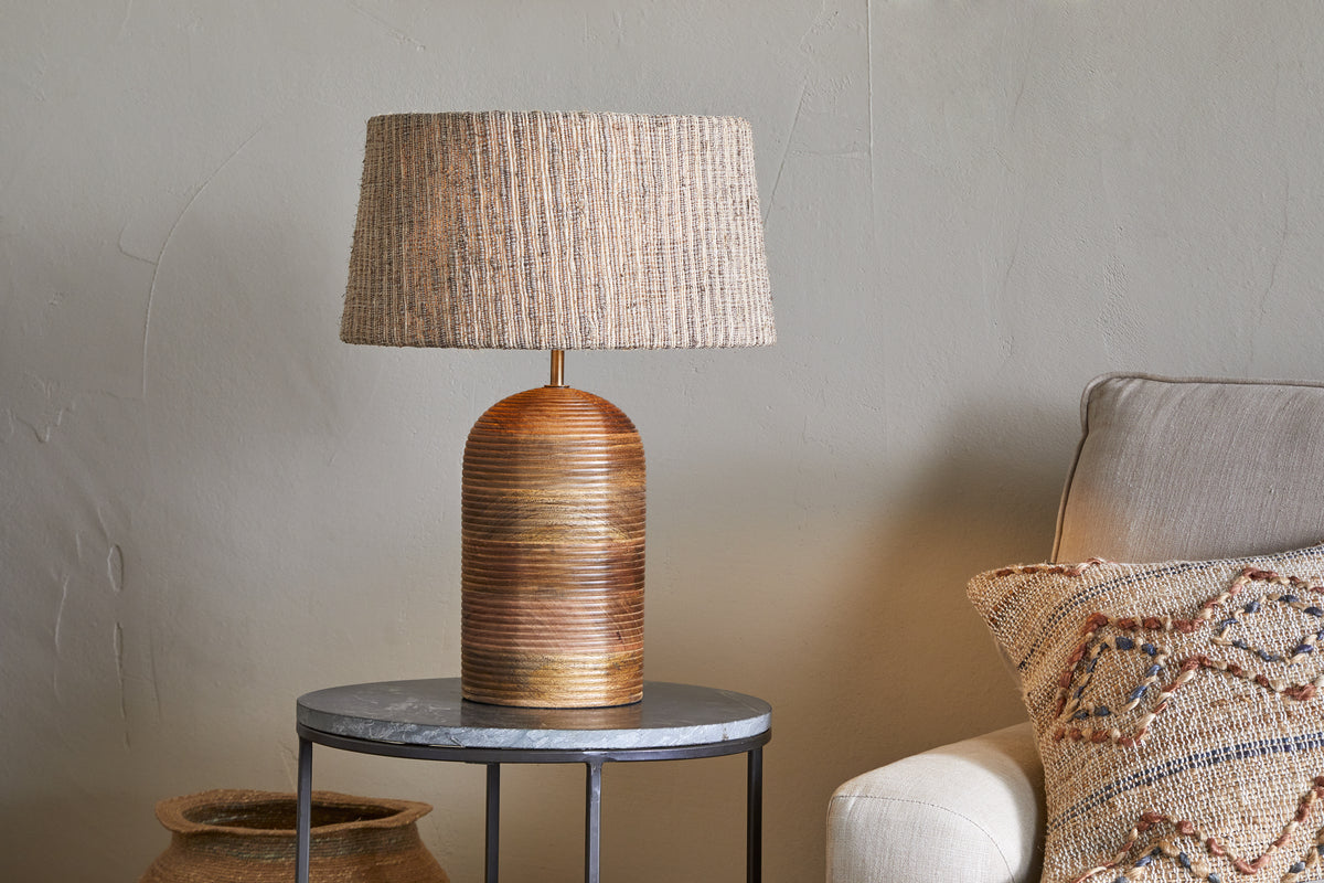 The nkuku Agua Grooved Table Lamp in natural, featuring a textured woven shade, sits on a round gray side table next to a light beige sofa with a patterned cushion. A woven basket is partially visible on the floor behind it.