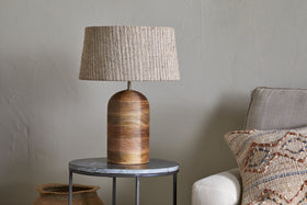 The nkuku Agua Grooved Table Lamp - Natural, featuring a textured woven beige shade, sits on a round dark metal side table with a stone top. Nearby are part of a beige sofa with a patterned pillow and a woven basket by the light gray wall.