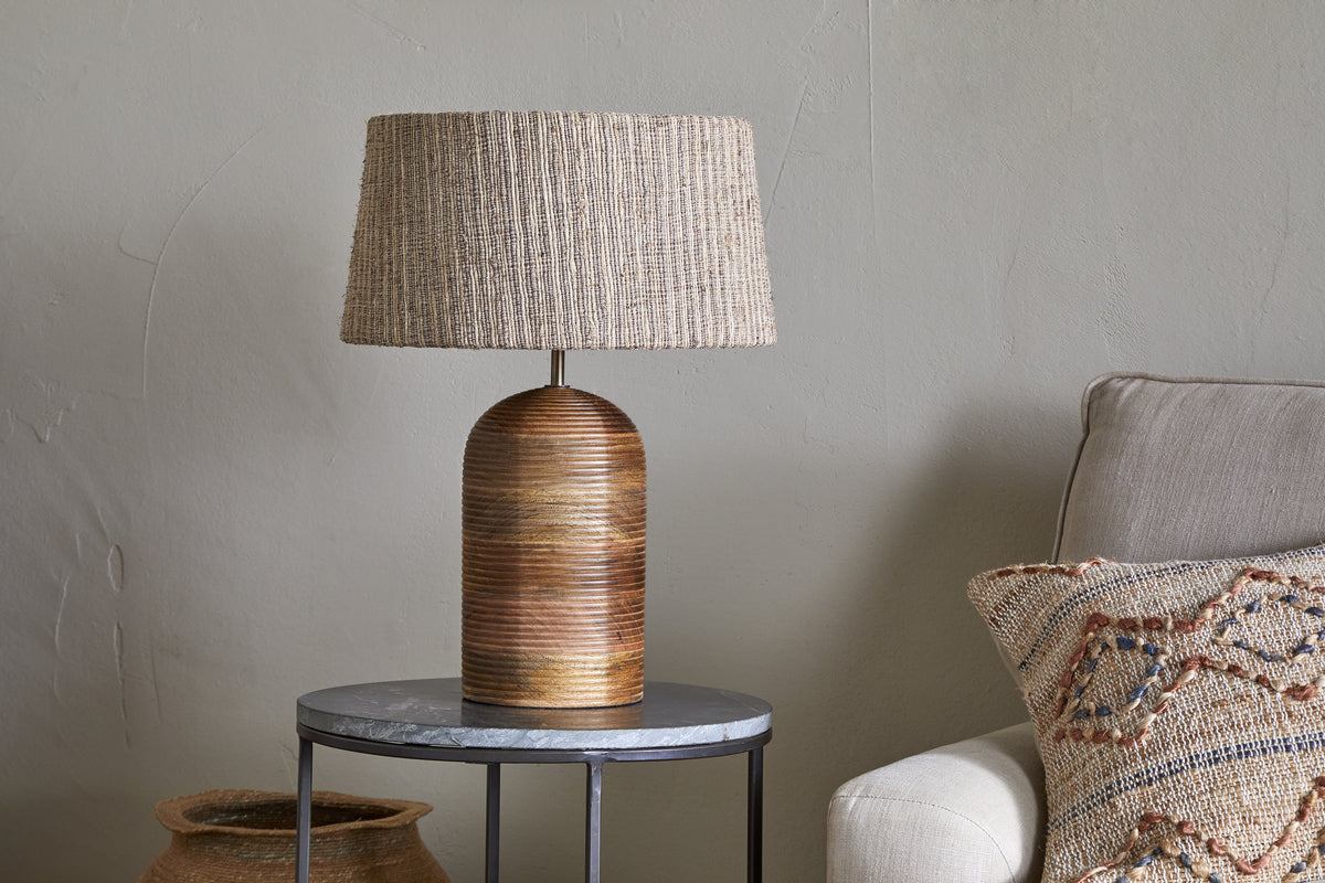 The nkuku Agua Grooved Table Lamp - Natural, featuring a textured woven beige shade, sits on a round dark metal side table with a stone top. Nearby are part of a beige sofa with a patterned pillow and a woven basket by the light gray wall.