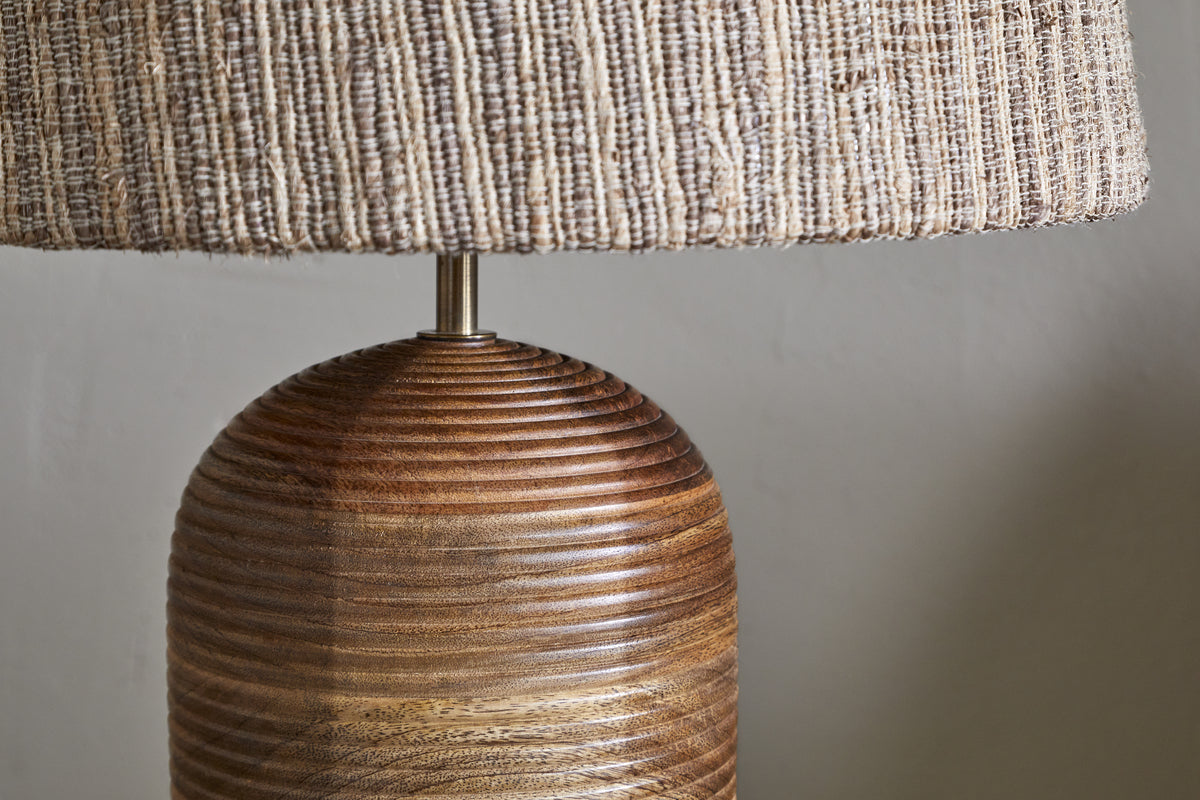 Close-up of the nkuku Agua Grooved Table Lamp - Natural, showcasing its hand-grooved mango wood base with a glossy finish and a textured, woven shade in earthy tones, set against a plain, light-colored wall.