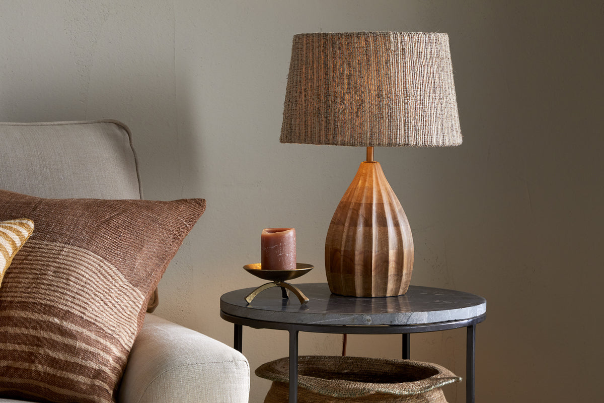 A cozy living room with a beige sofa and brown patterned pillows. Next to it, a round black side table holds nkuku's Awaa Mango Wood Bedside Table Lamp - Natural with a woven shade, plus a lit brown candle on a gold dish. A basket rests below.