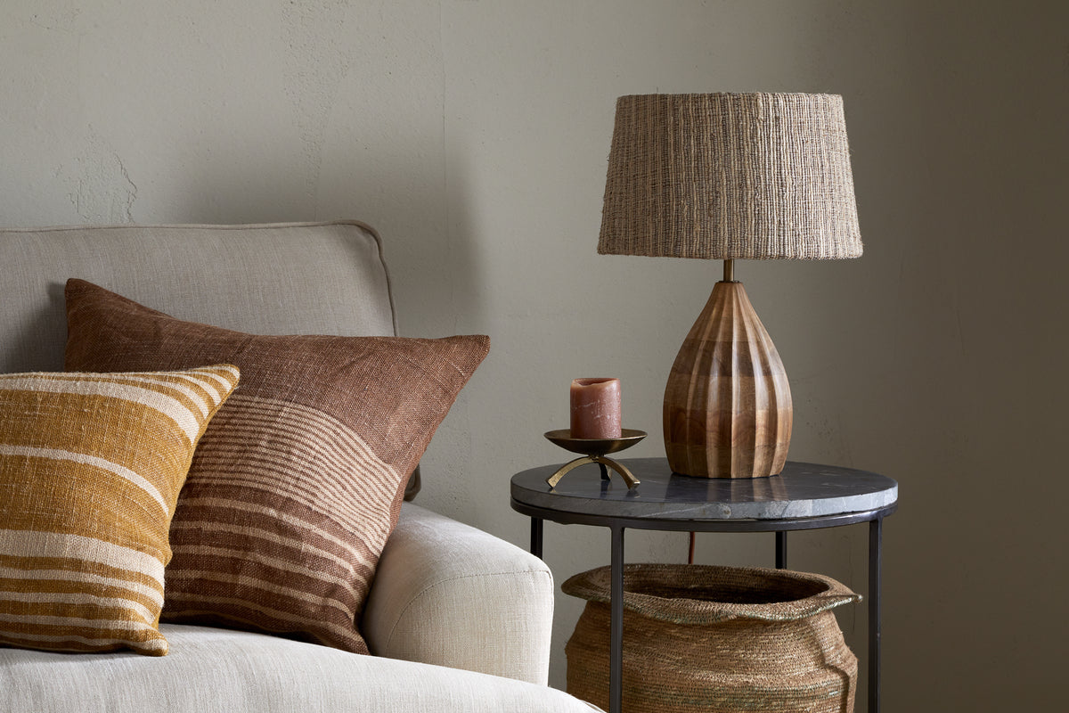 A cozy corner features a beige sofa with two textured brown and mustard pillows. Next to it, the nkuku Awaa Mango Wood Bedside Table Lamp in Natural sits on a round side table with a brown candle and woven basket below, set against a neutral wall.