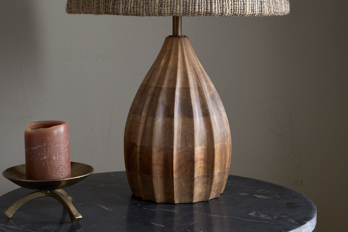 The nkuku Awaa Mango Wood Bedside Table Lamp, featuring a fluted mango wood base and woven shade, is displayed on a dark marble surface next to a brown candle in a brass holder against a plain light-colored wall.