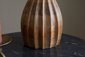The Awaa Mango Wood Bedside Table Lamp by nkuku, in natural finish, features vertical faceted grooves and warm wood grain. It sits on a dark marble-like surface with a brown candle on a brass plate to the left, set against a neutral background.