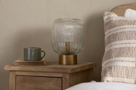 A wooden nightstand holds the nkuku Rizal Fluted Glass Bedside Table Lamp in antique brass & clear, a green mug, and a closed book. Striped beige and white pillow and white bedding are seen near a light-colored wall.