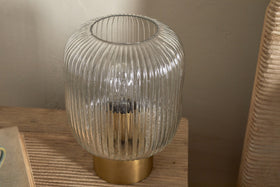 The Rizal Fluted Glass Bedside Table Lamp by nkuku, featuring a cylindrical clear glass design and antique brass base, rests on a wooden surface with sunlight casting soft shadows beside a beige wall and woven chair.